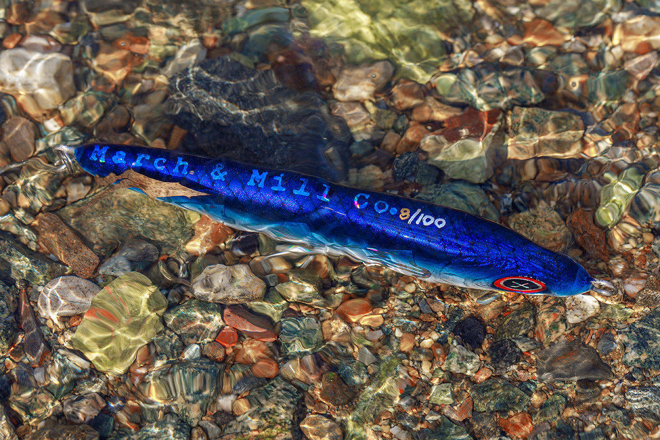 Blue fishing lure with 'March & Hall Co' branding on a bed of colorful pebbles.