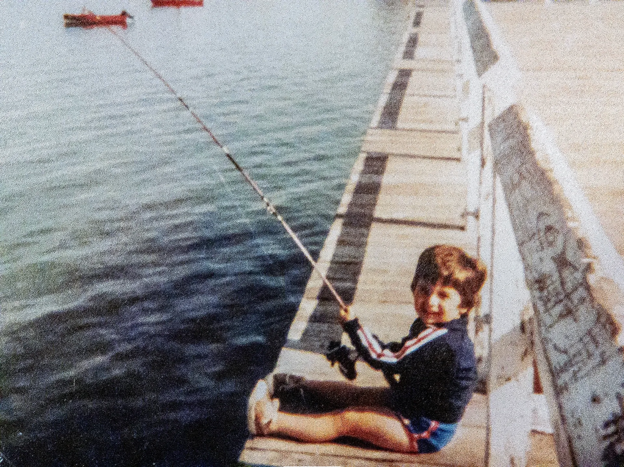 Child sitting on a wooden dock fishing in a body of water with a boat in the background.