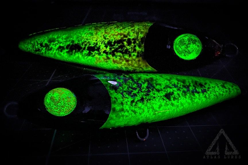 Two glowing green fishing lures on a dark surface with a brand logo in the corner.