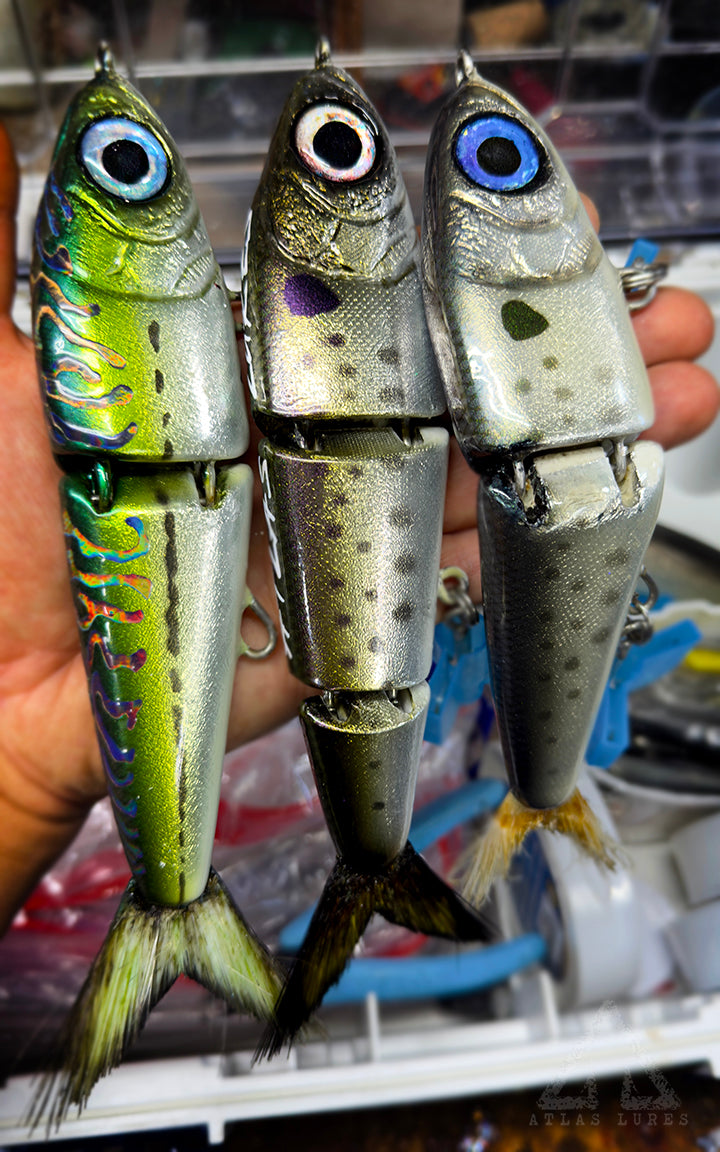 Three fishing lures resembling silver mackerel held by a hand with a blurred background.